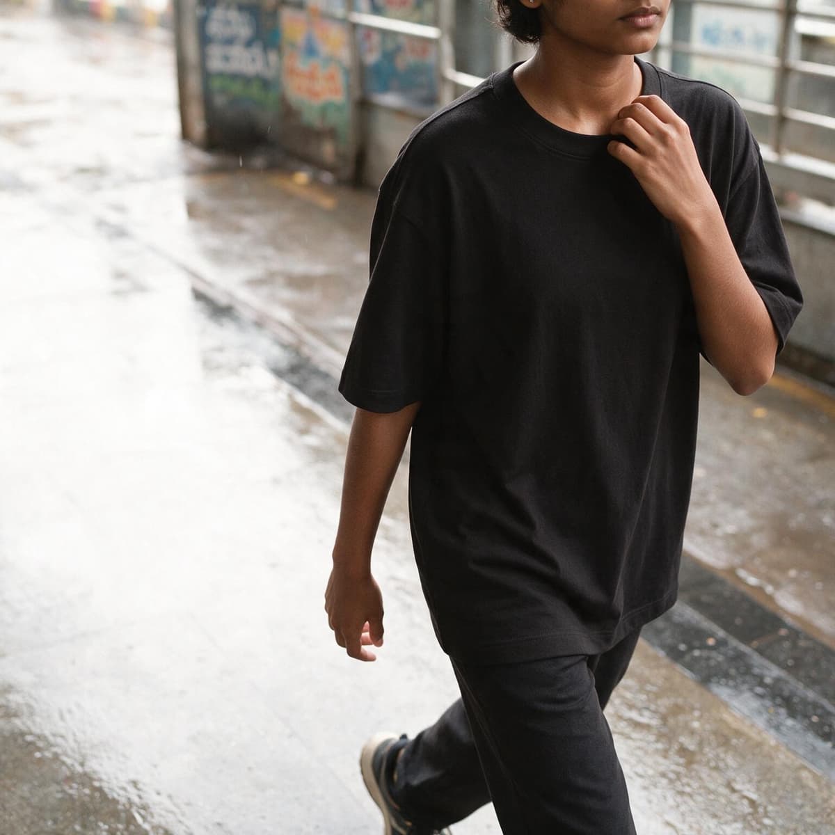 Person wearing a black oversized classic round neck tee walking on a wet city pavement after rain.